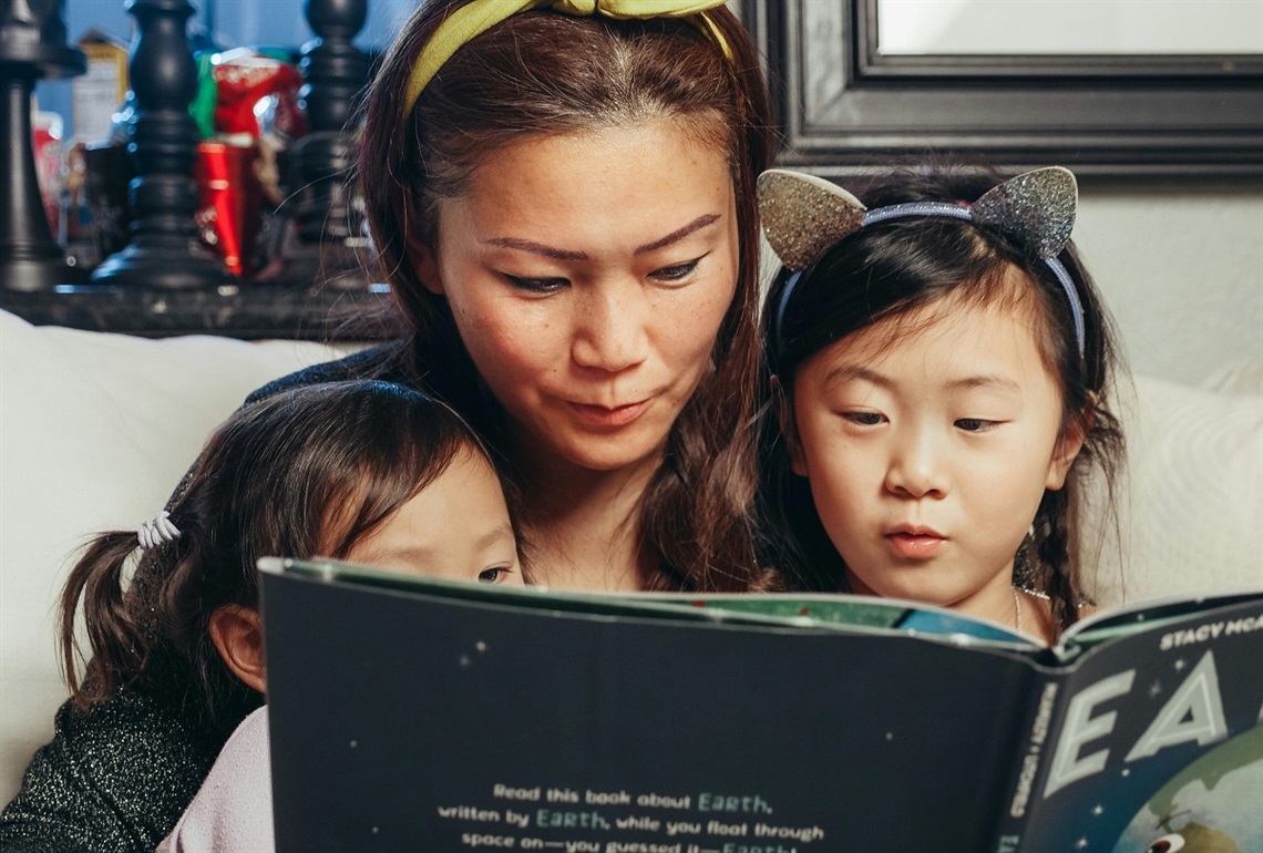 A woman reads a book with her two young daughters on her lap.