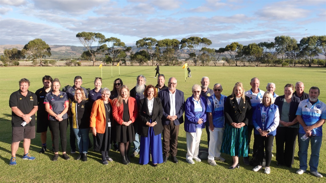 A large group of people, including uniformed sports club members, smile for the camera on a grass soccer pitch at the launch of upgraded Aldinga Sports Park facilities.