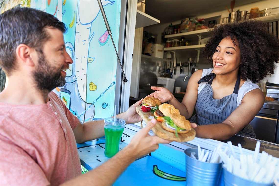 A smiling curly-haired woman passes a bearded man three burgers on a wooden platter from a food truck window.