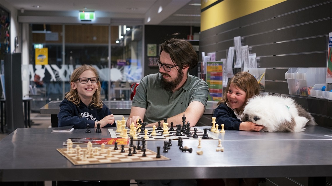 Two young current Children's University participants sit alongside an older previous participant with two chessboards on a table in the foreground.