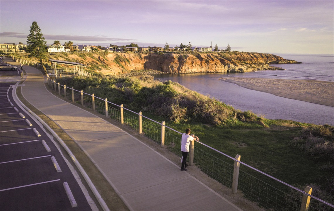 A person leans over the railing along a clifftop path at Port Noarlunga overlooking the ocean and cliffs in afternoon sunlight.