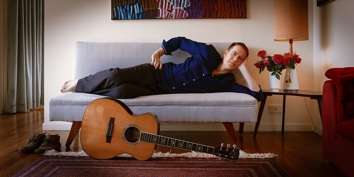 Peformer Fred Smith lies horizontally on a couch on his side facing the camera wearing a blue shirt with a guitar on the floor in front of him.