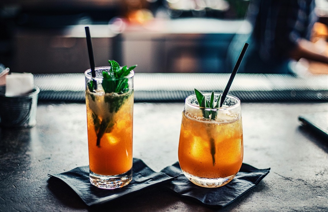 Two glasses with an orange-brown liquid inside and adorned with green leaves sit on a bar.