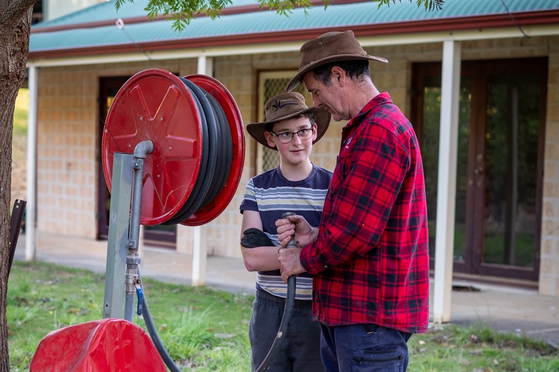 A father and son in brown wide-brimmed hats inspect a fire hose alongside a red reel at their rural property.