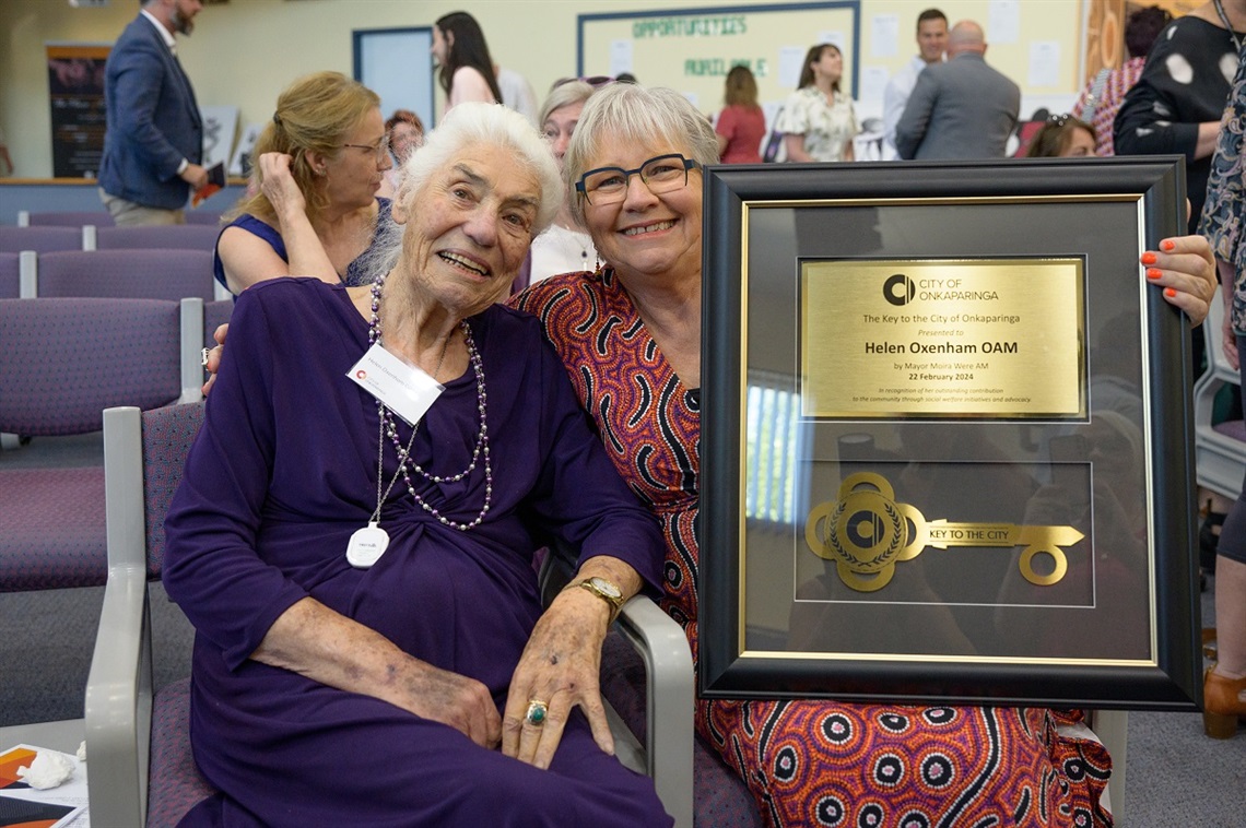 A smiling Helen Oxenham OAM in a purple dress sits alongside Onkaparinga Mayor Moira Were, who holds a framed Key to the City.