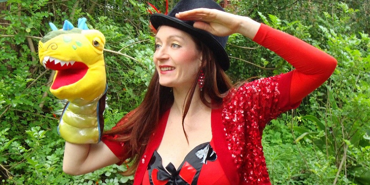 Jesstar the puppeteer in a red outfit and black hat smiles with a yellow dinosaur-like puppet.