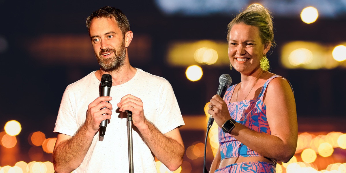 Comedians SA’s Kel Balnaves and NT’s Amy Hetherington standing side-by-side smiling on stage with microphones.
