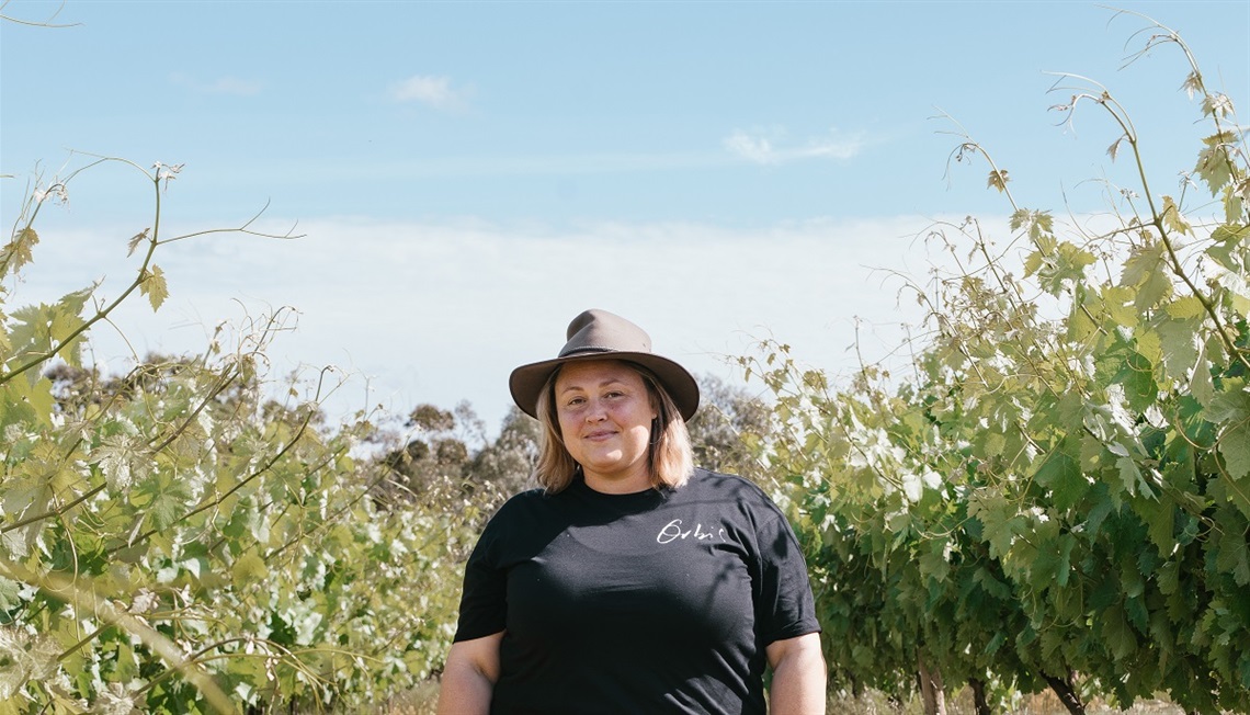 Lauren Langfield from Orbis Wines stands in a black T-shirt and brown wide-brimmed hat among the vineyards.