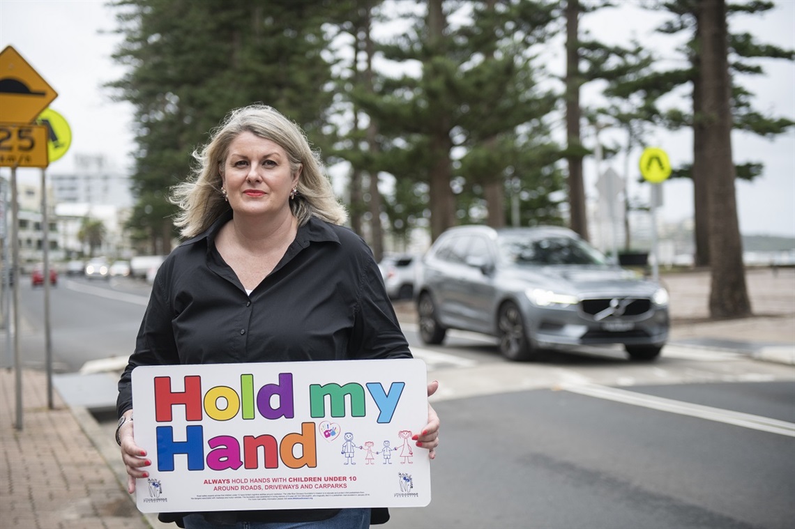 Michelle McLaughlin holds a colourful sign that reads 'Hold my Hand' alongside a tree-lined road with a car in the background.