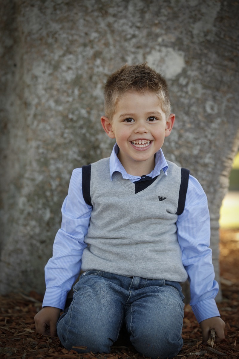 Tom McLaughlin, 4, smiles and kneels in the leaves alongside a tree trunk wearing jeans, a blue shirt and a grey vest.