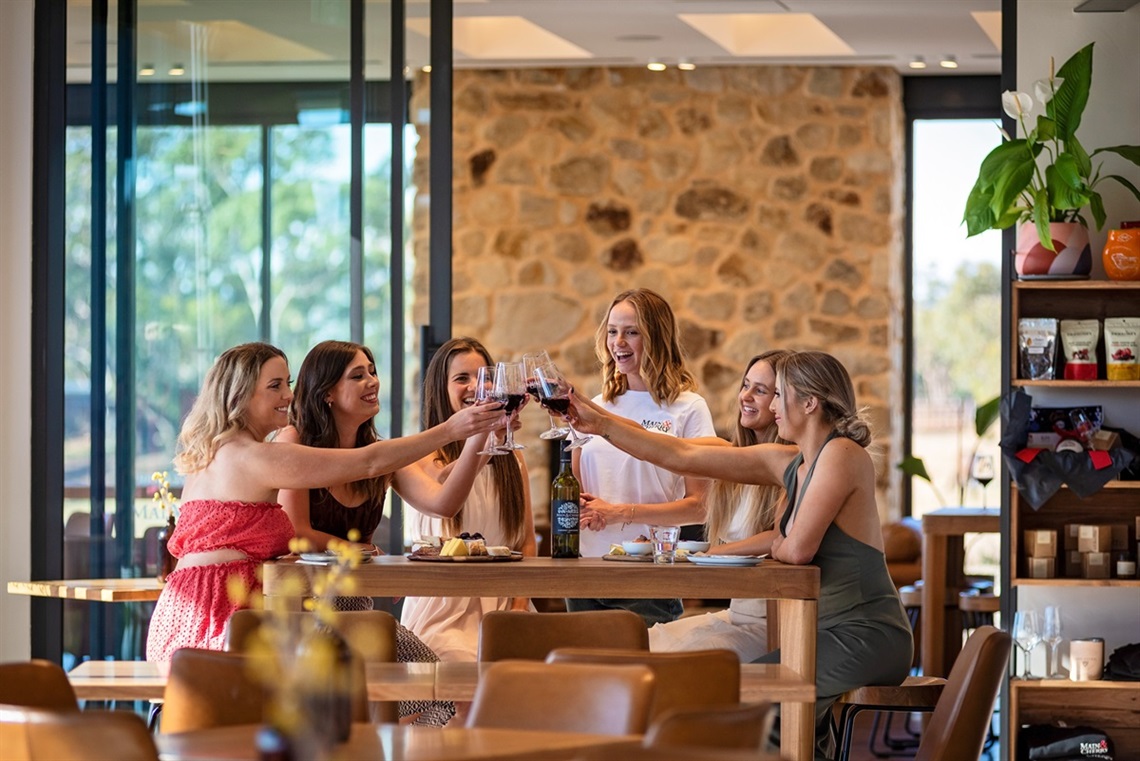 A group of smiling women at a cellar door table toast their glasses of red wine together.