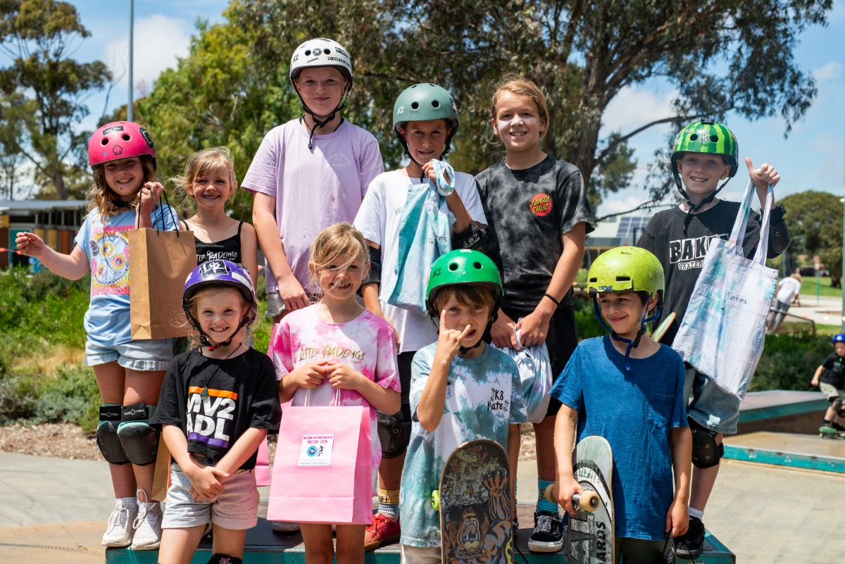 A group of young skateboarders in colourful T-shirts smile at a skate park.