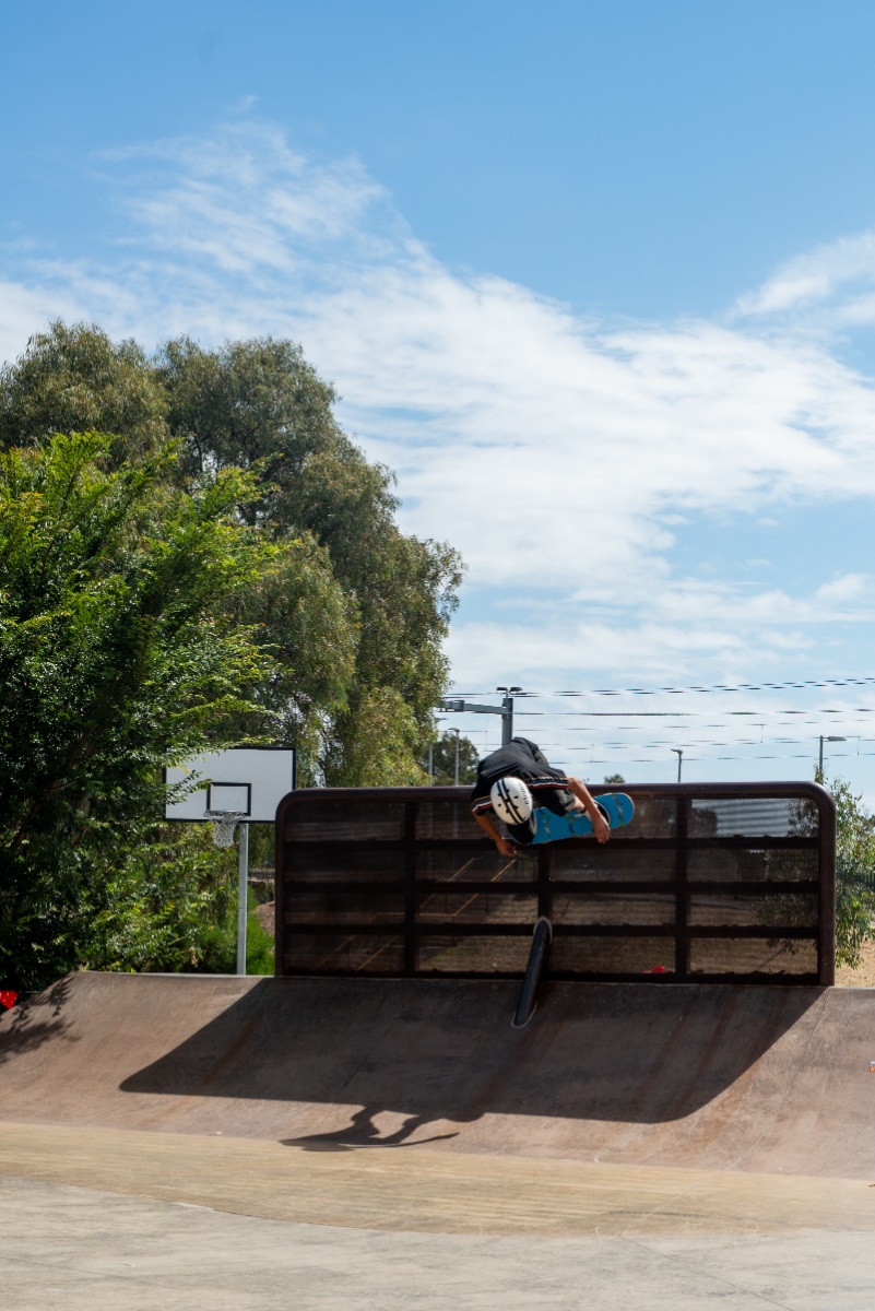 A skateboarder skates high along a walled section at a skate park with blue sky, a basketball ring and trees behind him.