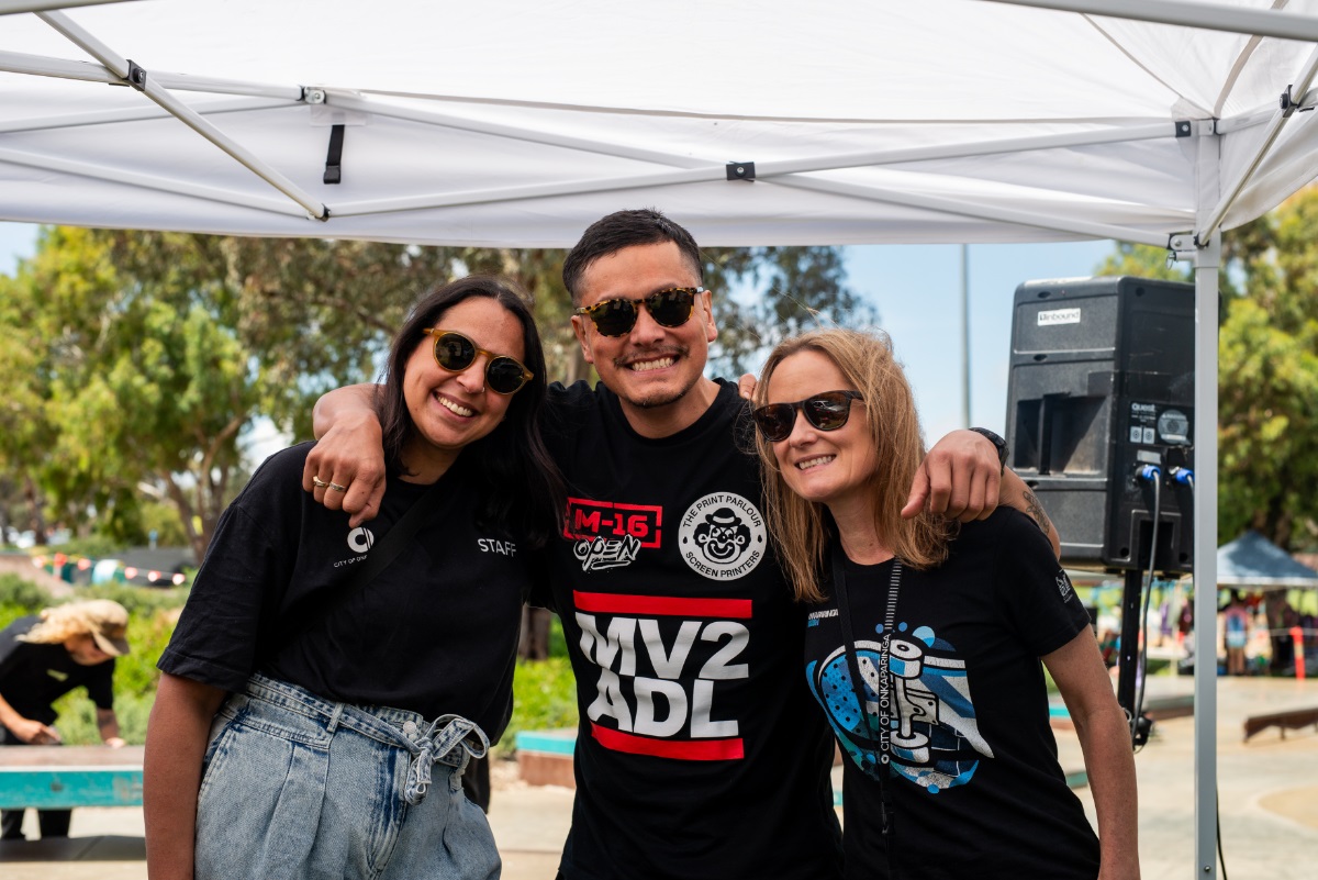 A smiling man wearing a black MV2 ADL T-shirt has his arms around two smiling women in black T-shirts. All are wearing sunglasses.