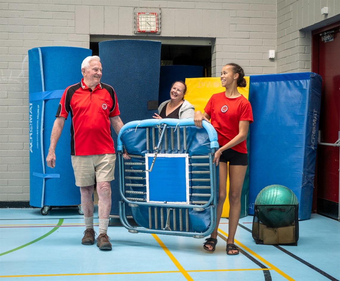 Neville Gibbs carries a trampoline with a young gymnast, with another smiling Morphett Vale Youth Club member behind them.