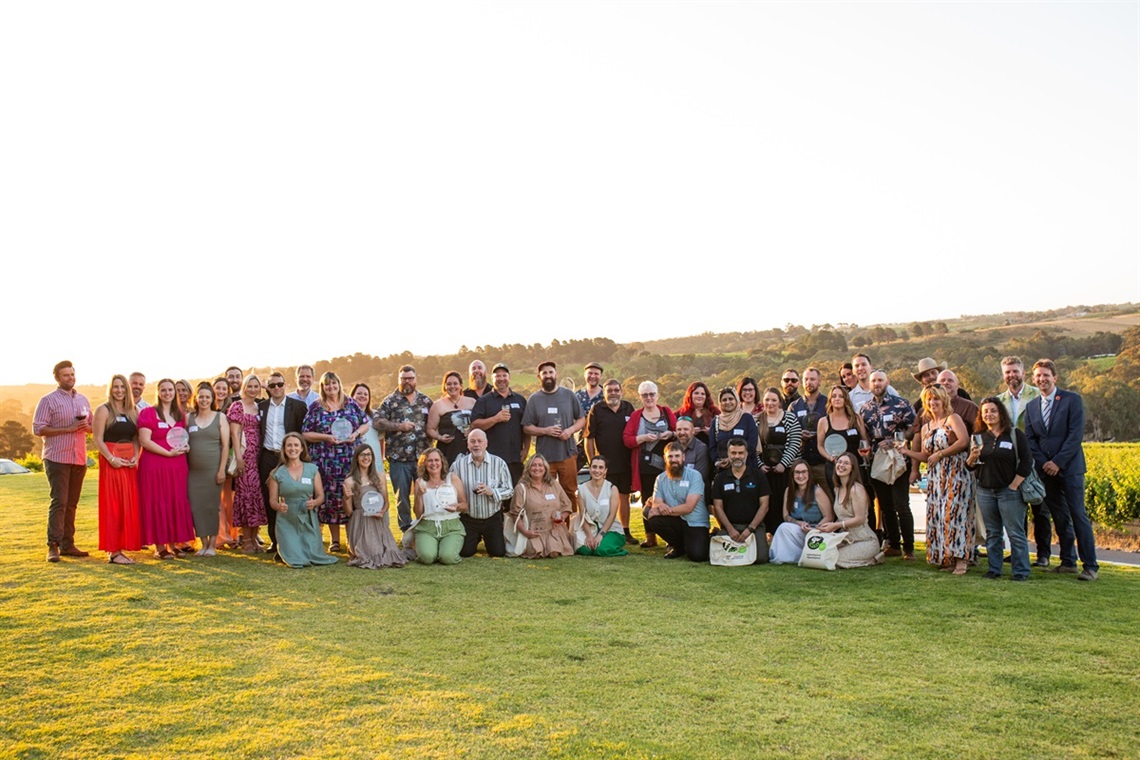 A group shot of the many businesses to receive Onkaparinga Business Awards smiling with their awards on grass with the sun setting behind them.