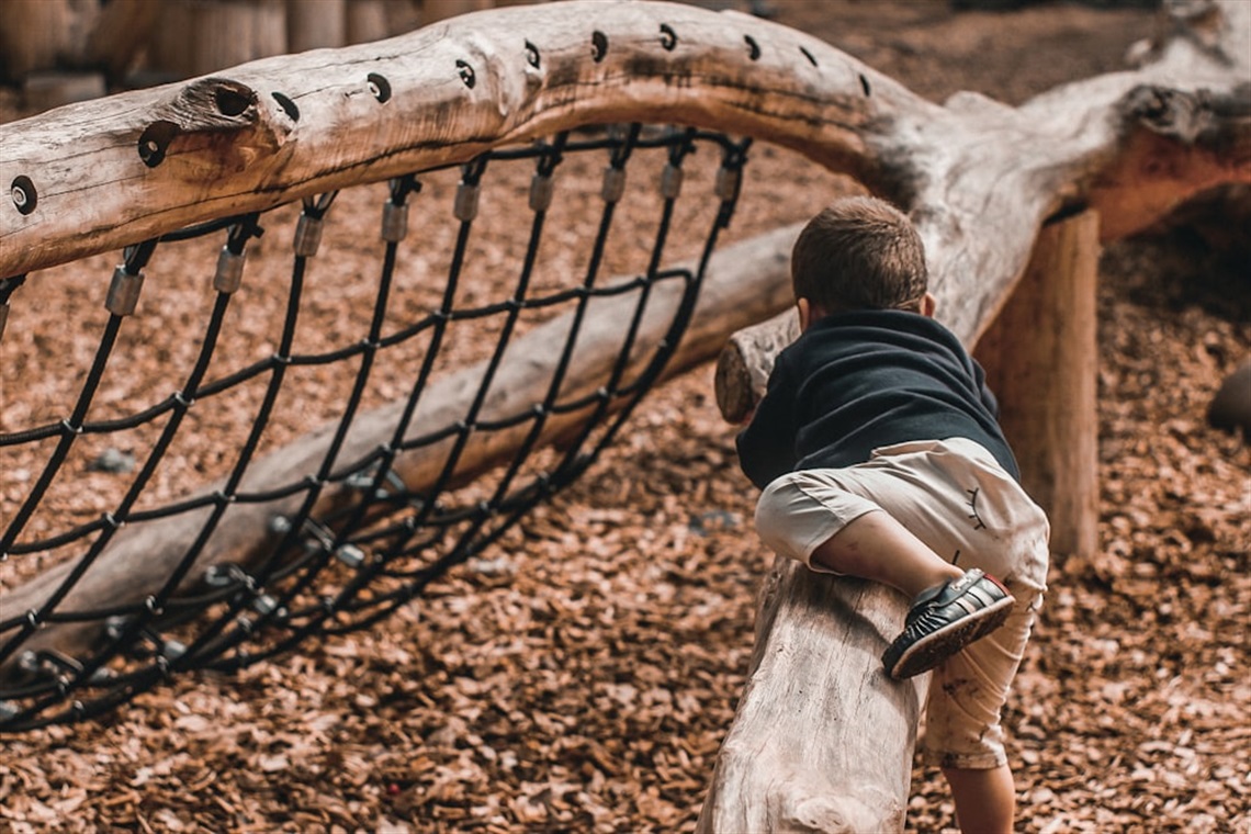 A young boy climbs over a wooden post at a nature playground with barkchips beneath him and a play net ahead of him.