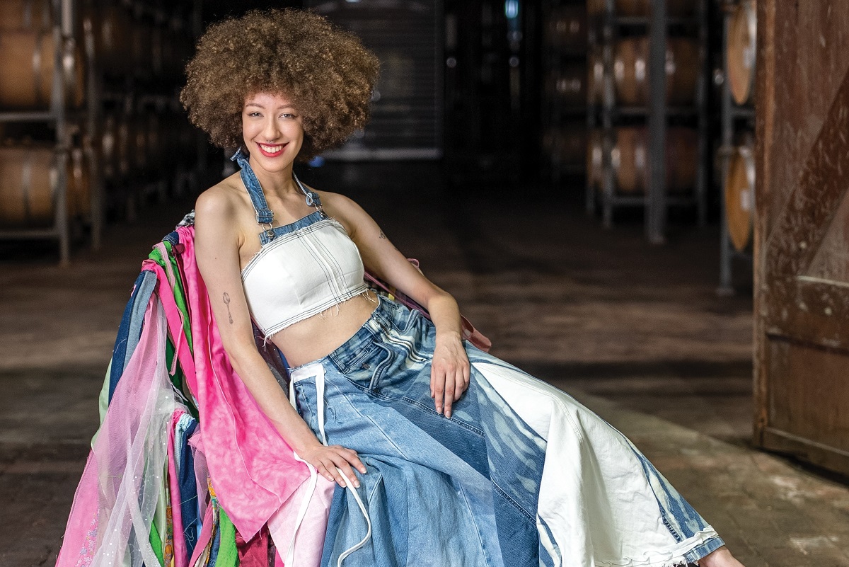 A smiling, curly-haired model reclines in a chair wearing a recycled denim-heavy outfit atop colourful recycled materials.