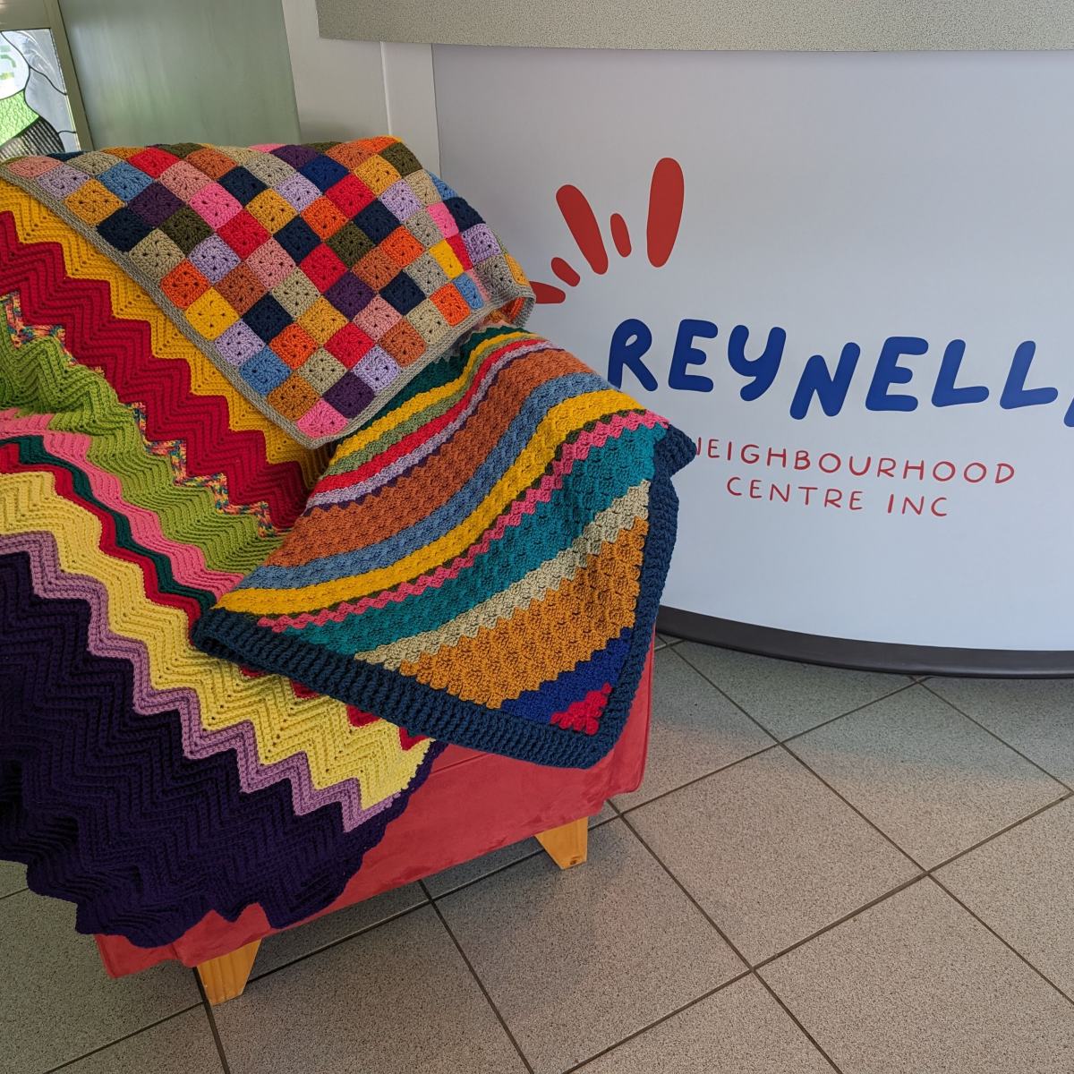 Colourful quilts are displayed on a single-seater couch with a Reynella Neighbourhood Centre Inc sign in the background.