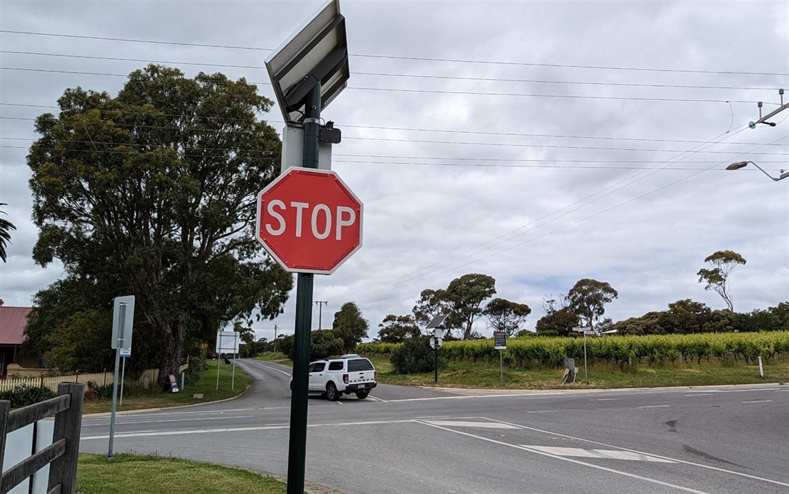 A photo of an RJAWS stop sign equipped with active warning technology at  a McLaren Vale intersection with vineyards and a white ute in the background.