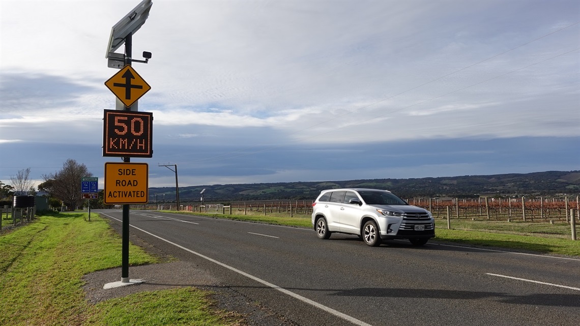 A flashing warning sign is activated as a silver SUV drives past it on a rural McLaren Vale road with vineyards in the background.