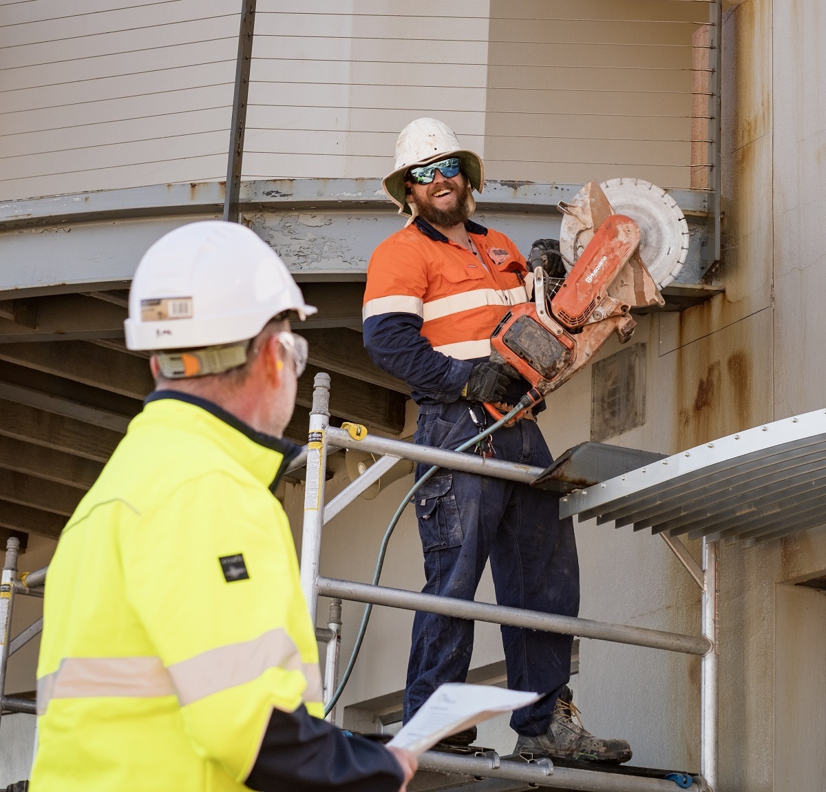 RMK Building Group's Robbie looks up at subcontractor Alex, who smiles down at him while handling a circular saw.