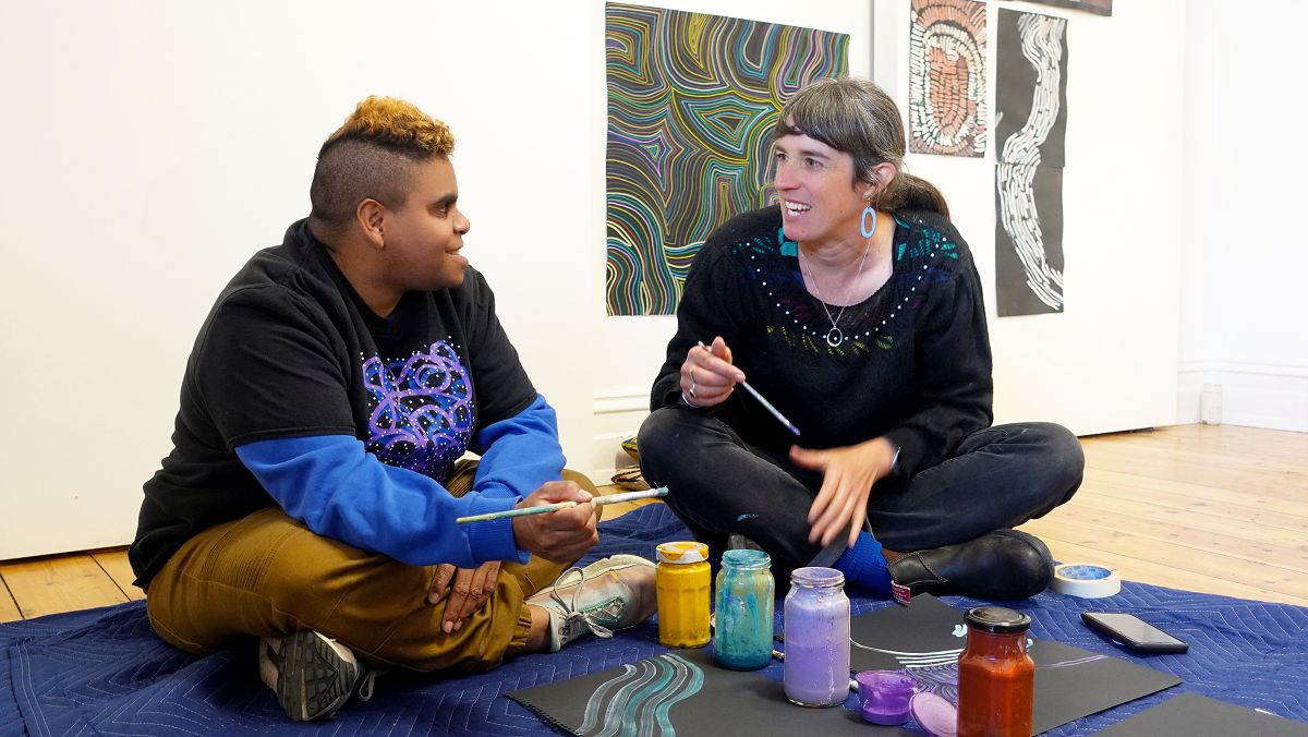 Artists Jackie Saunders and Laura Wills sit cross-legged at their studio and smile at one another with paintbrushes in their hands.