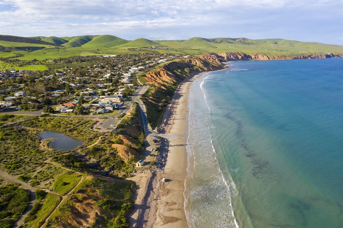 An aerial photo of Sellicks Beach, featuring the township, green rolling hills, ochre cliffs and flat blue ocean.