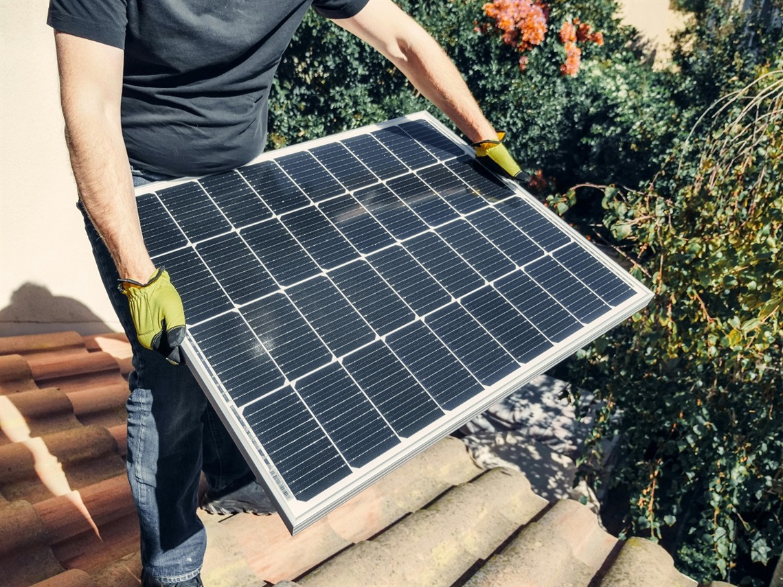 A tradesperson holds a solar panel with yellow gloves atop the roof of a house.