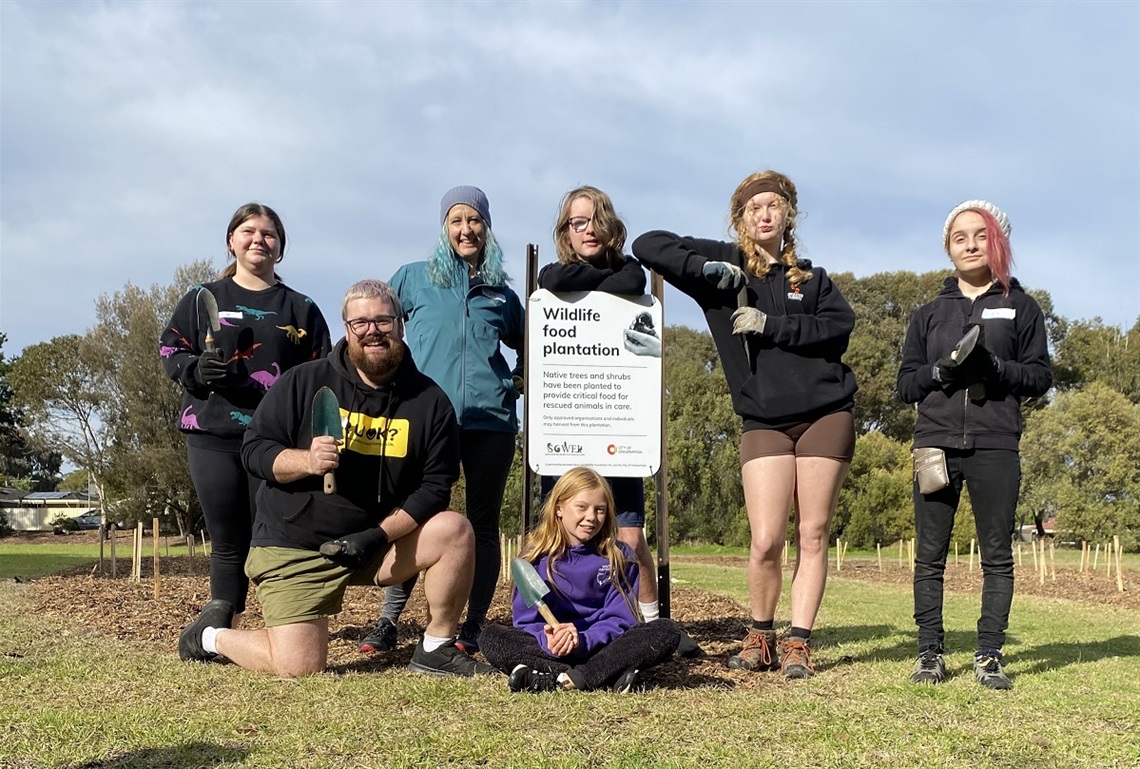 A group of smiling council staff and young volunteers smile with gardening trowels at a Happy Valley reserve.
