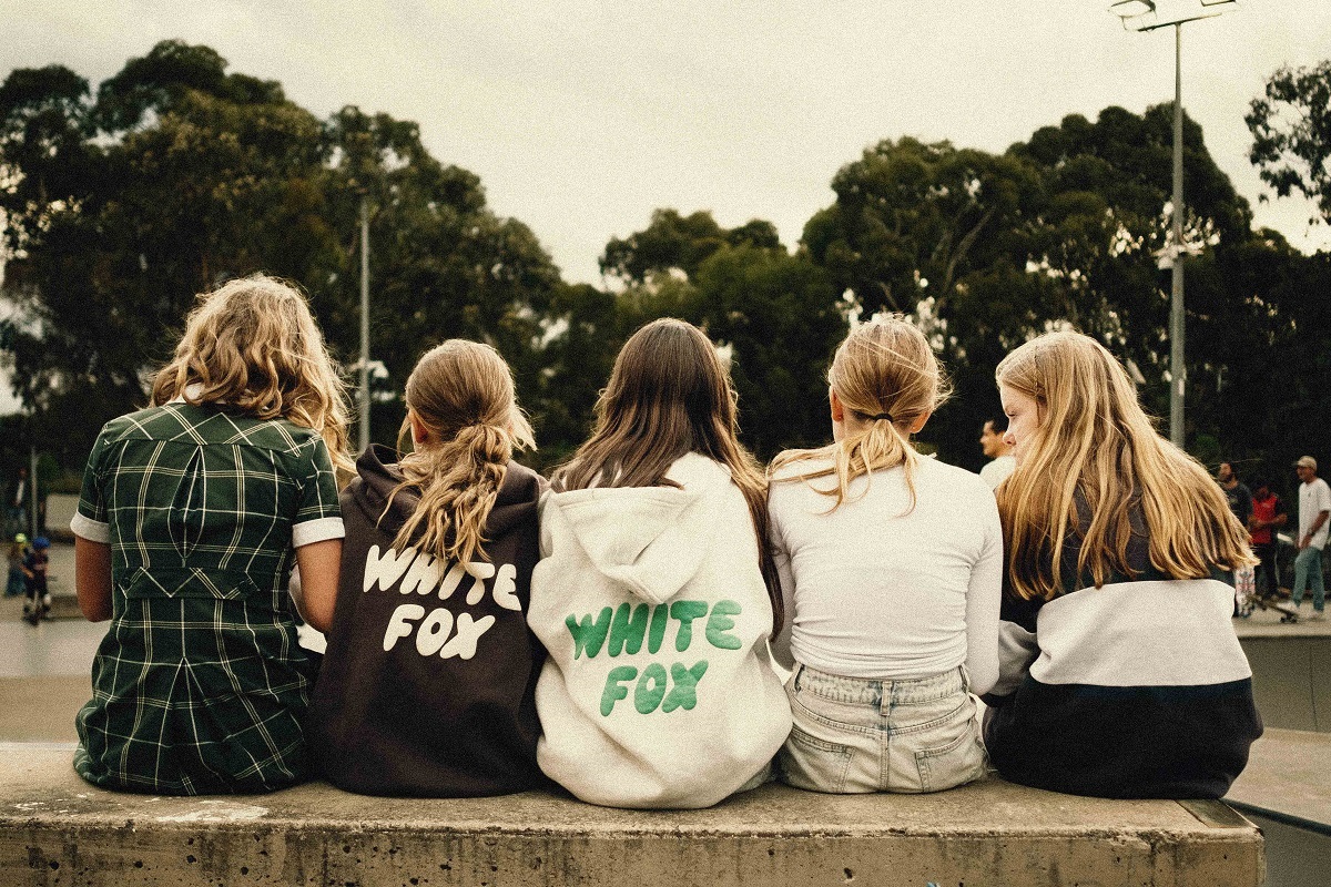 A group of young people with long hair sit alongside one another at a skate park, photographed from behind.