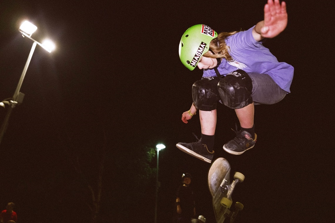 A skateboarder in a purple T-shirt and green helmet performs a flip trick on a skateboard at night.