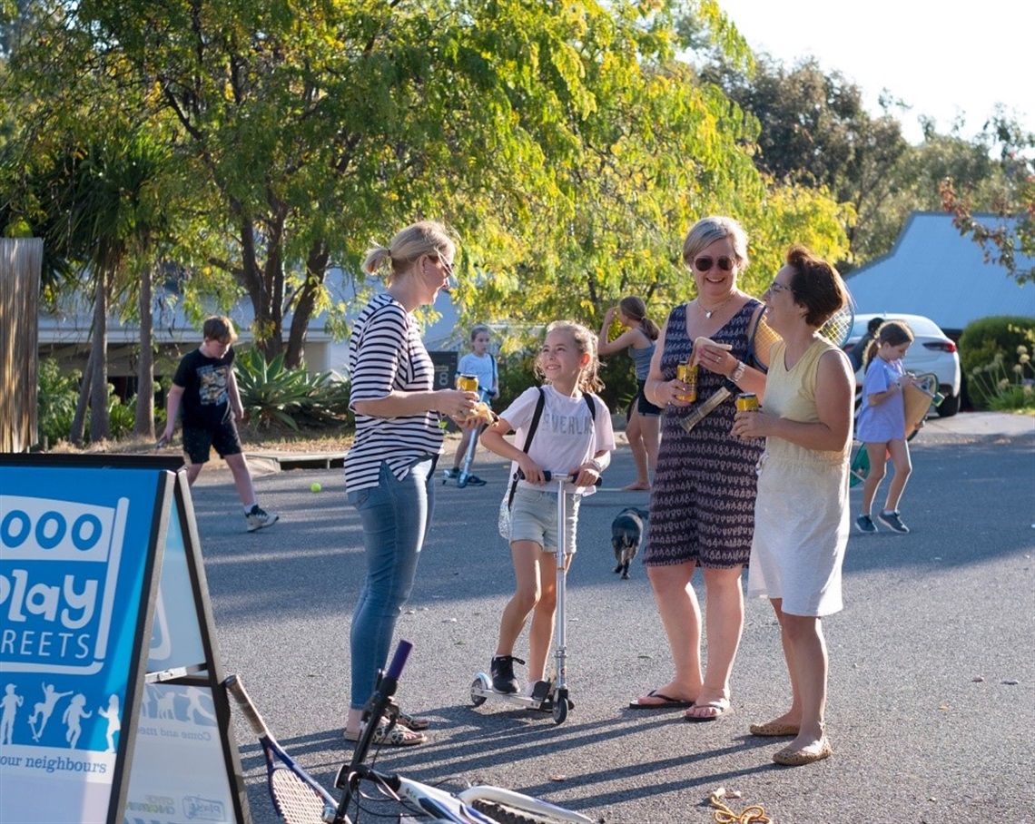 Three smiling women and a girl on a scooter chat on a suburban street with leafy trees in the background.