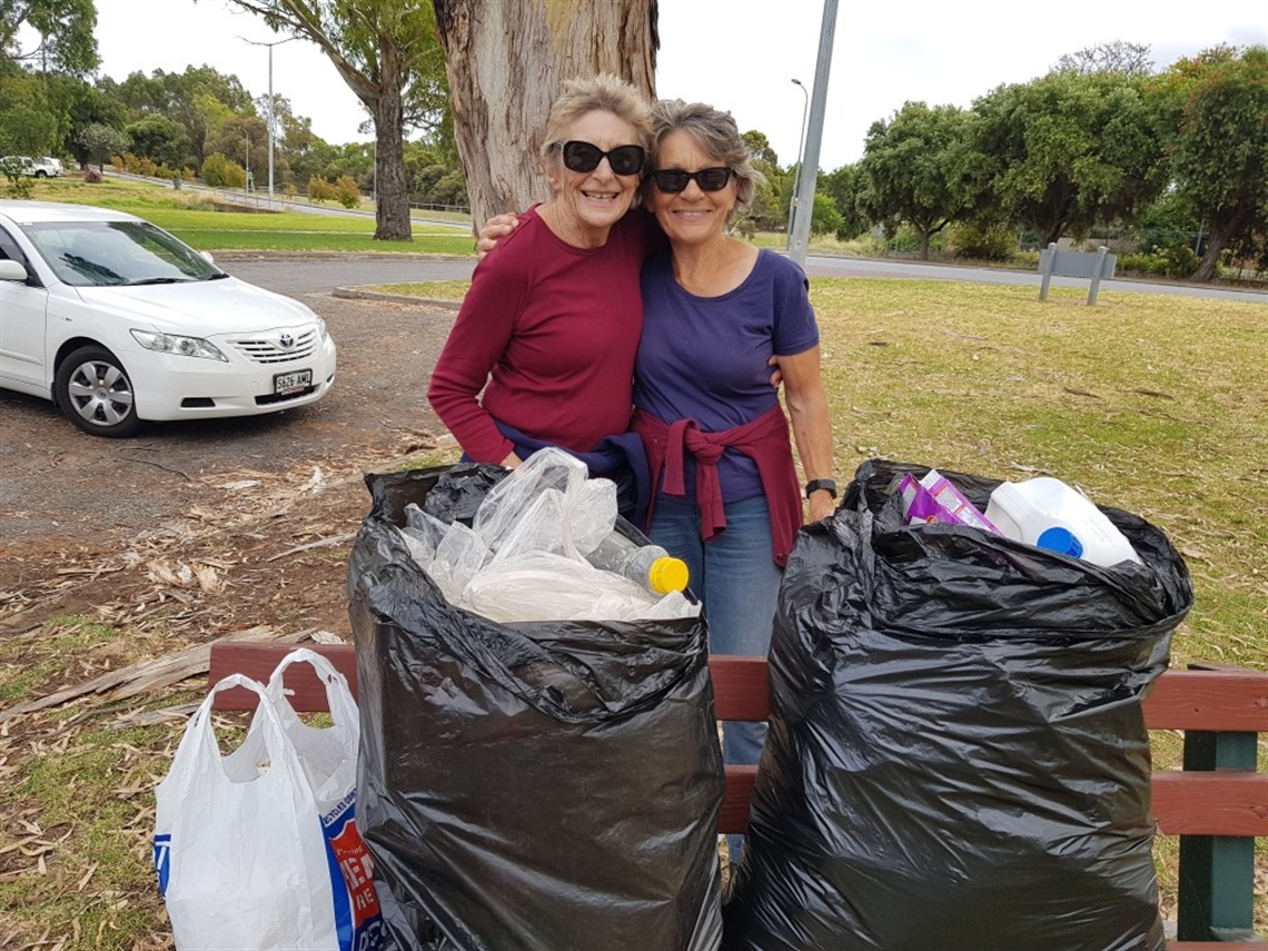 A photo of two smiling friends with their arms around one another with two large bags of rubbish in front of them.