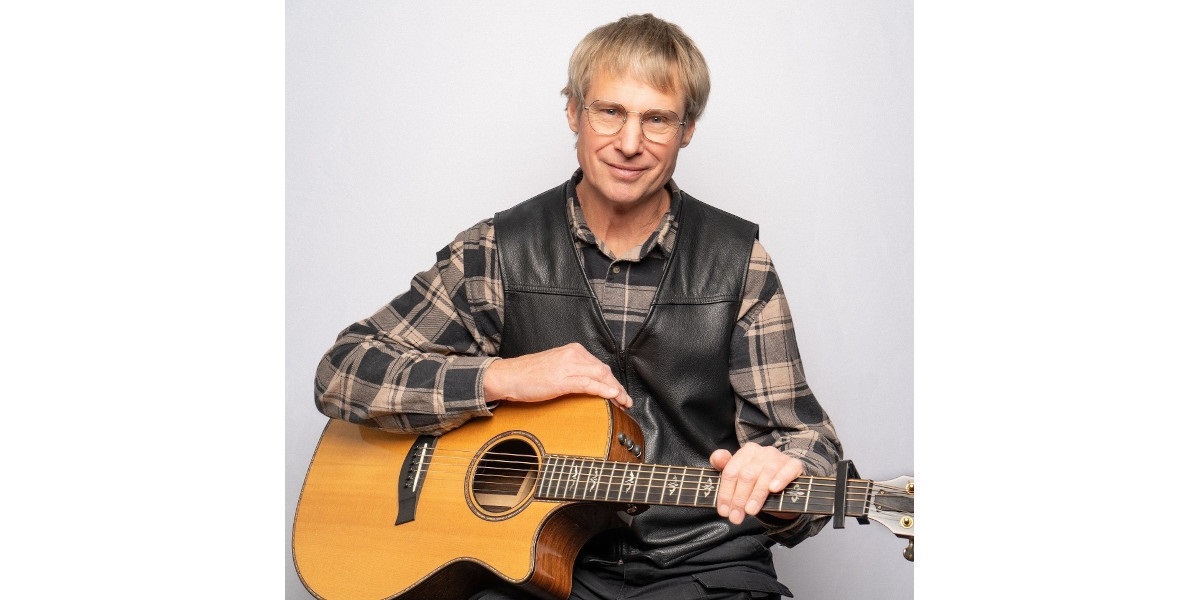 John Raymond dressed as John Denver in glasses, a black vest and chequered shirt holding a guitar.