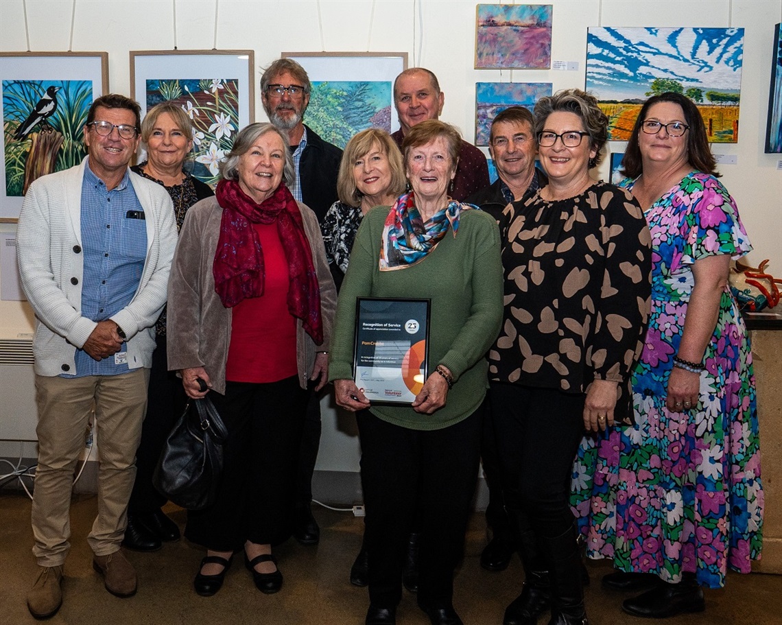 A group of smiling volunteers, a woman in front holding a volunteering certificate from the City of Onkaparinga, stand in front of some paintings.