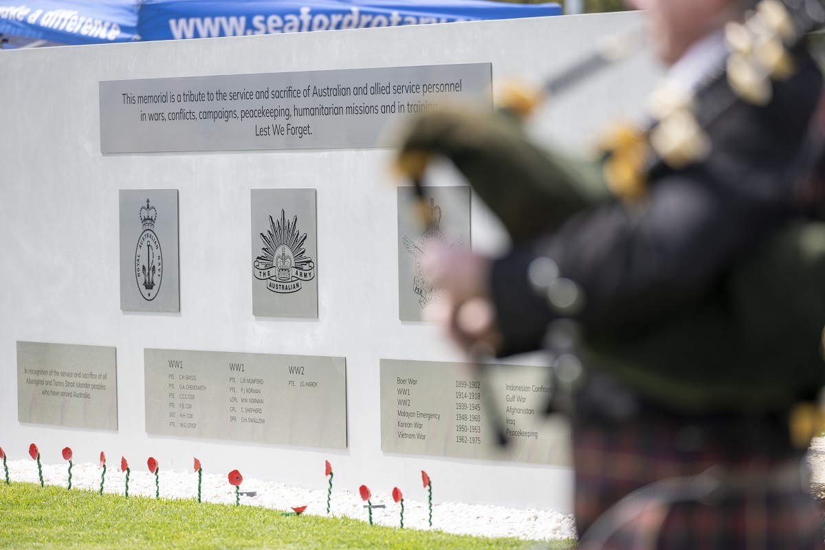 A plaque-filled wall at the new memorial with a blurred bagpipe player in the foreground.