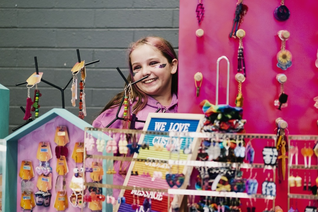 A girl with pink-streaked hair smiles from behind a stall selling jewellery.