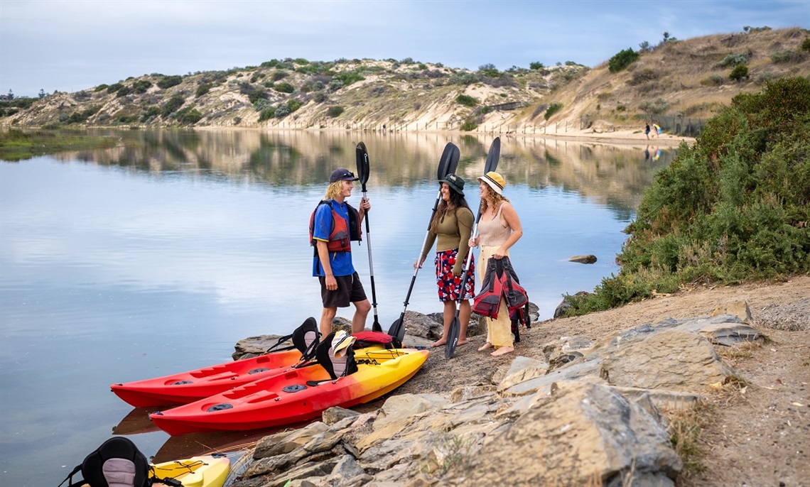 Three kayakers smile at one another on the banks of the Onkaparinga River in Port Noarlunga alongside two red-and-yellow kayaks while holding paddles.