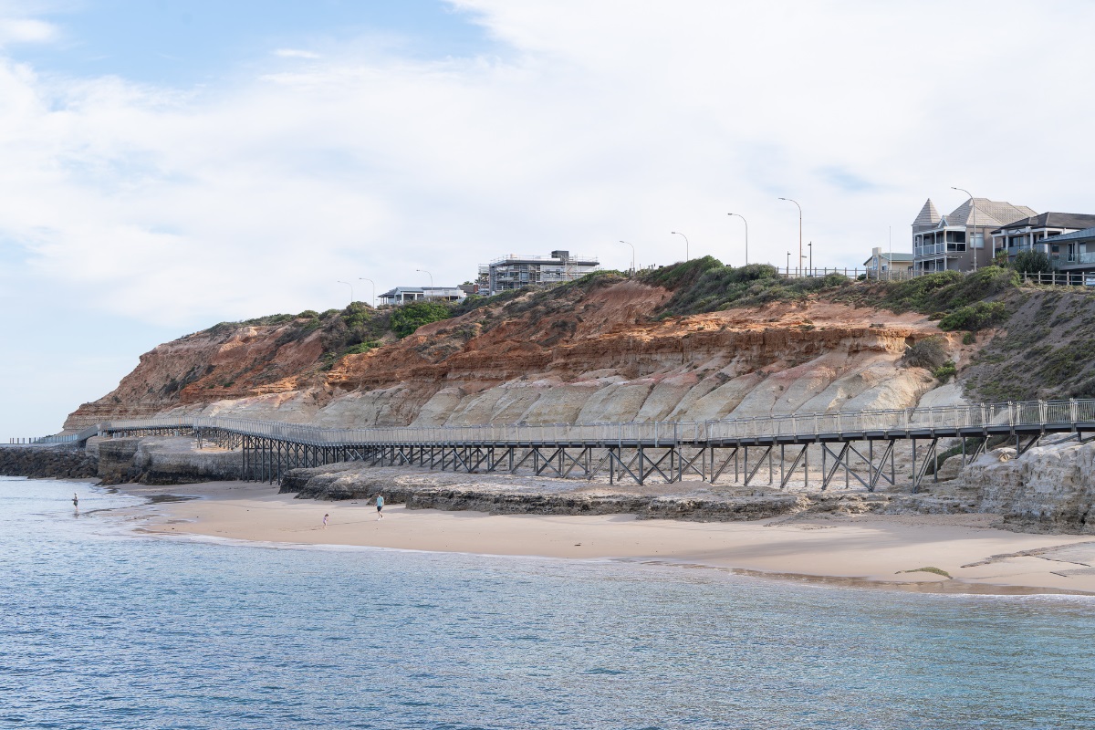 A view from the water of the Witton Bluff Base Trail boardwalk with orange cliffs in the background and a parent and child playing on the sand.