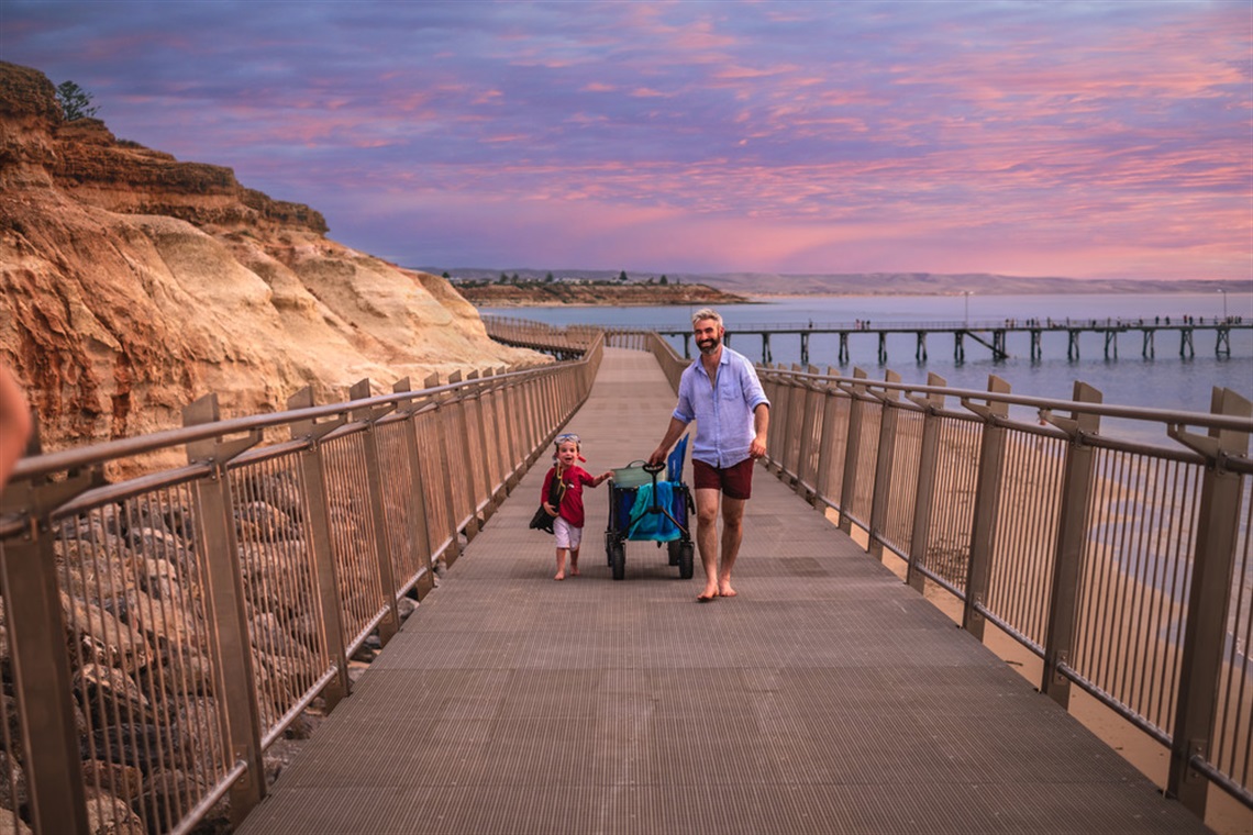 A young boy and his father walk along the Witton Bluff Base Trail boardwalk pulling a beach wagon behind them, with the evening sky beginning to turn pink and purple behind the Port Noarlunga jetty.