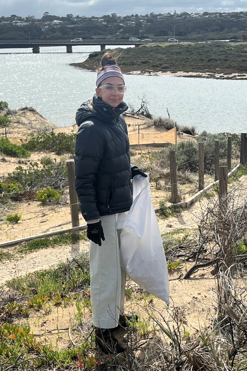 A woman wearing a black jacket and clear protective glasses stands in some sand dunes with a white bag and the Onkaparinga River behind her.