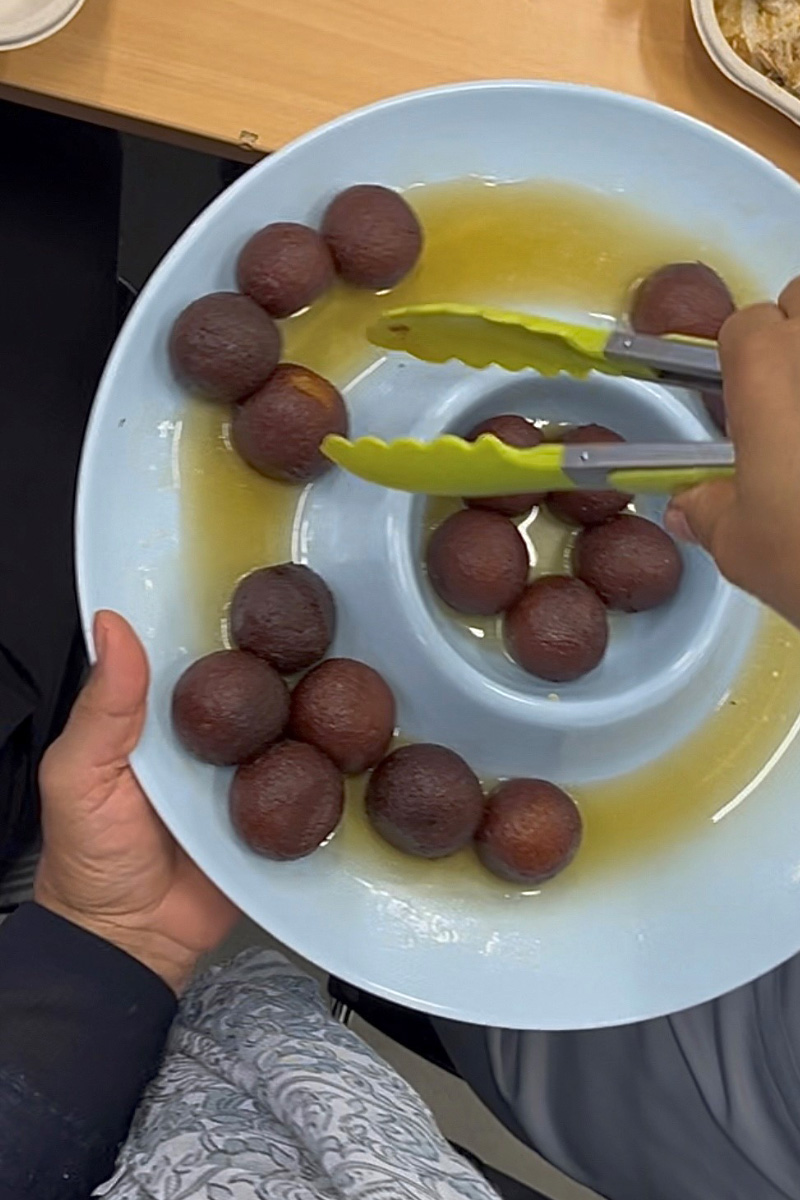 A photo of Gulab Jamun balls on a round plate with a pair of tongs in someone's hands hovering above it.