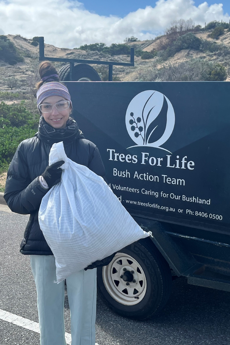A smiling woman in a black jacket carrying a white bag stands in front of a trailer with Trees for Life signage on it.