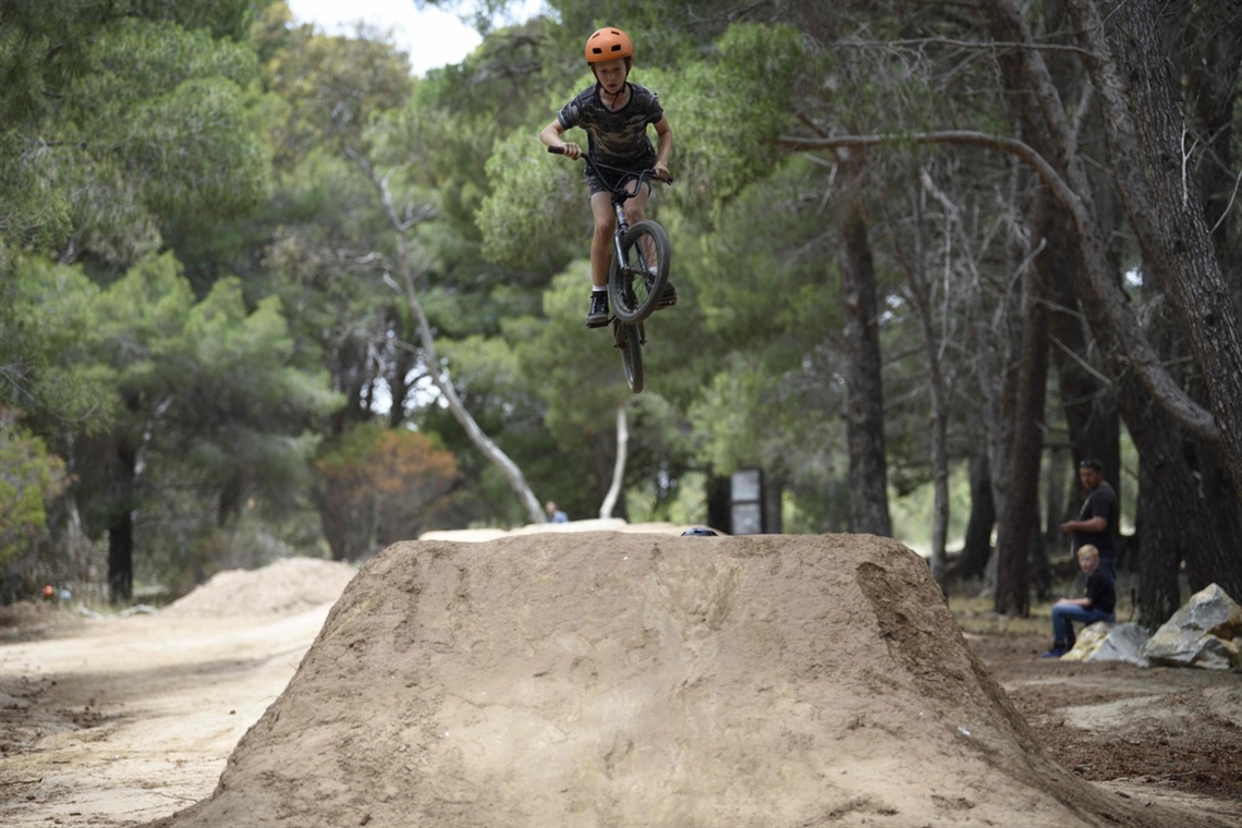 A young BMX rider soars above some dirt jumps with trees alongside and behind them.