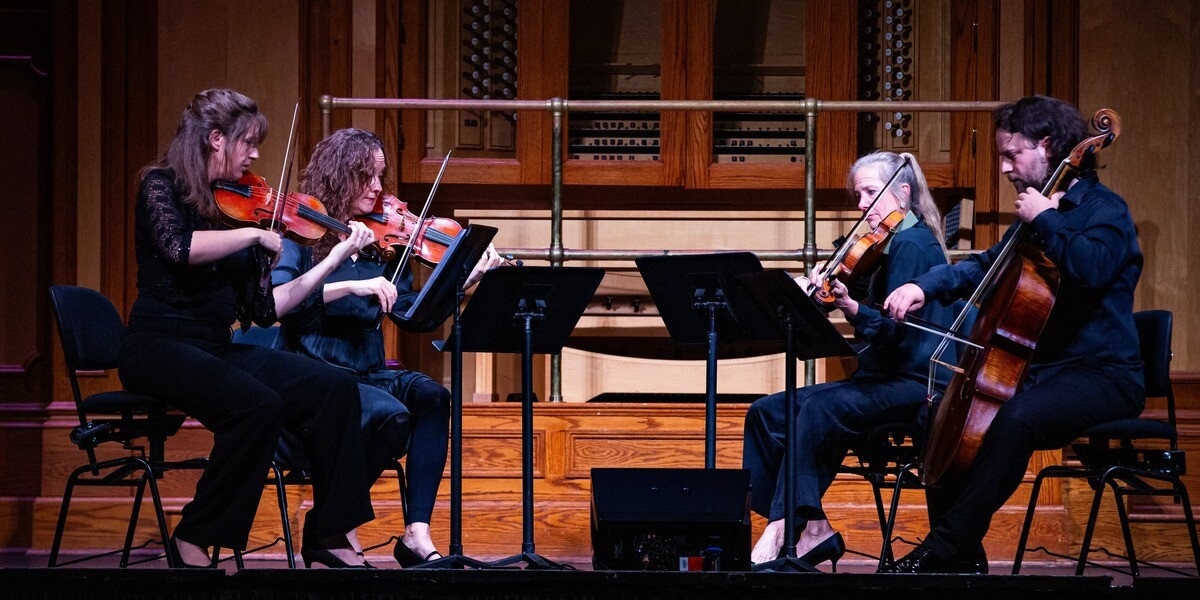 Four musicians play their string instruments while looking at music sheets on stage.