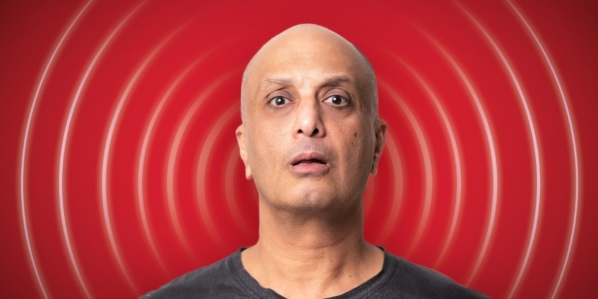 Comedian, Akmal, stares down the barrel of the camera framed by a red background similar to a Loony Tunes introductio.