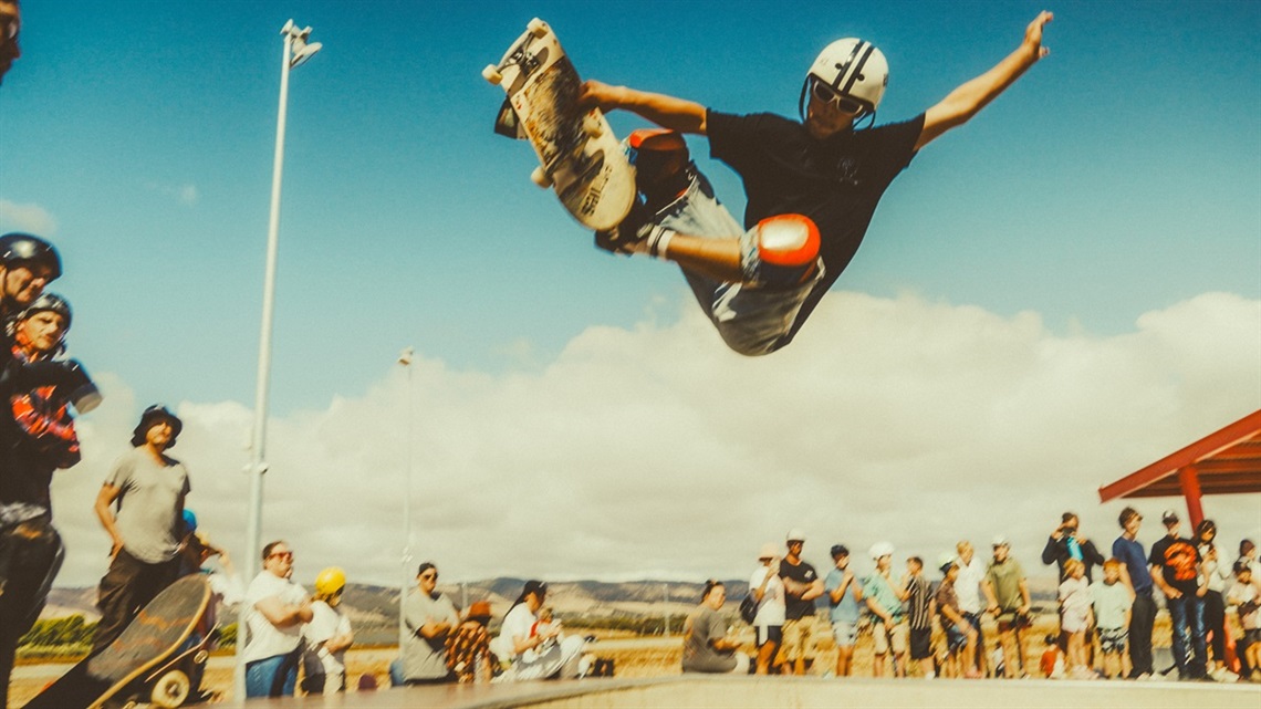 A helmeted skateboarder grabs his board as he launches an air out of the Aldinga skate park with onlookers standing behind him.