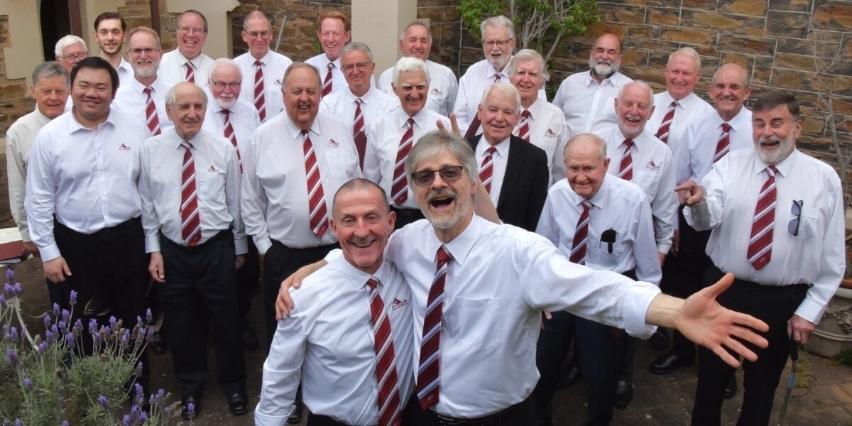 A large group of men wearing white shirts with red-and-white ties sing while looking at the camera.