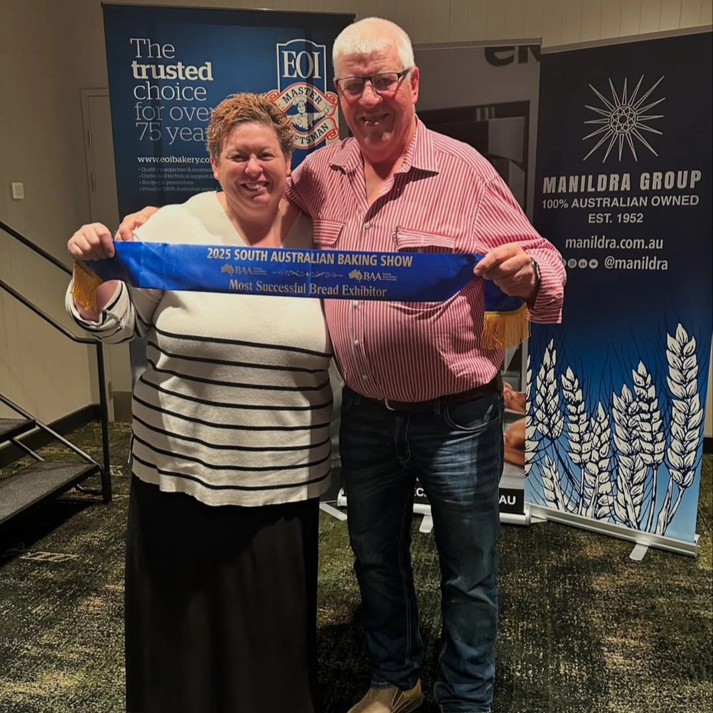 A man and woman hold up a blue sash that names them winners of the Most Successful Bread Exhibitor.